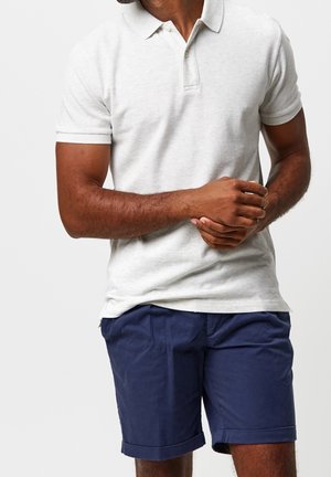 Mężczyzna w jasnoszarej, krótkorękawowej koszulce polo i granatowych szortach z podwiniętymi nogawkami, stoi ze splecionymi rękami przed sobą.