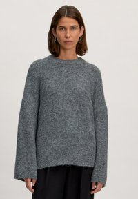 Grau gestrickter Pullover mit lockerem Schnitt, rundem Ausschnitt und langen Ärmeln. Strukturierter Stoff und ungesäumtes Detail am Saum. Über einer schwarzen Hose getragen.