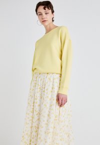 Gul genser med avslappet passform og rund hals, parret med en hvit plisert skjørt med små gule blomstermønstre.