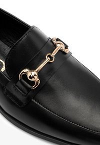 Zwarte leren instapper met een ronde neus, voorzien van een goudkleurige metalen schakelaccent over de bovendkant en gedetailleerde stiksels langs de randen.