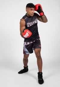 Zwarte mouwloze top en shorts met "Gorilla Wear" in vet roze, met een opvallend diagonaal gestreept patroon; rode bokshandschoenen; zwarte schoenen.