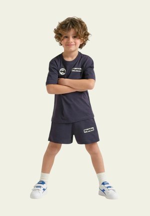 Ragazzo giovane con capelli ricci, in piedi con le braccia incrociate e un'espressione sicura, indossa abbigliamento sportivo navy Diadora e sneakers bianche con dettagli blu.