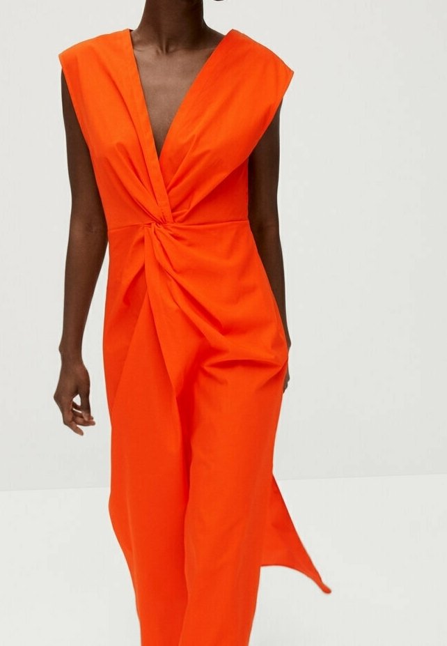 Robe sans manches orange vif avec un profond décolleté en V et un détail torsadé central, portée par une personne debout sur un fond clair uni.