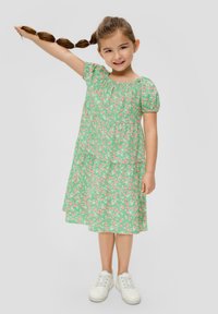 Robe fleurie verte avec des manches bouffantes, design à volants, ornée de fleurs roses sur un fond vert. Des baskets blanches complètent l'ensemble.