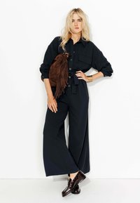 Chemise noire à boutons avec poches, associée à un pantalon large noir. Sac épaule frangé marron et chaussures sombres complètent le look.