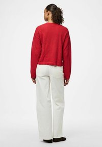Maglione rosso lavorato a maglia con un design testurizzato, lunghezza cropped e orlo a coste, abbinato a pantaloni beige a gamba larga e scarpe scure.