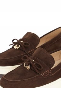 Brown suede loafers with a white sole, featuring a round gold charm and bow accent on the front, and a smooth interior lining.