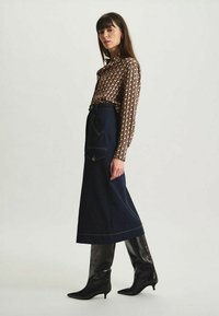 Longue jupe en denim avec une poche, associée à une chemise à boutons à motifs. Modèle avec des bottes à talons aiguilles noires et brillantes. Arrière-plan simple.