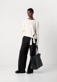Lichtbeige sweatshirt met een geknoopte zijde detail, gecombineerd met brede zwarte broeken en een zwarte tote tas. Glad materiaal en een relaxte pasvorm.