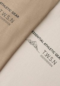 Beige og off-white stof med teksten "ESSENTIAL ATHLETIC GEAR T.W.S.N" og en enkel stregtegning af bjerge trykt i sort.