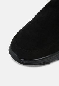 Svart mockasneaker med en slät yta, rundad form och gummisula med subtila sömnadsdetaljer längs kanten.