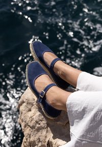 Espadrilles en daim bleu marine avec bout rond, bride à la cheville et détail en jute tissée sur la semelle extérieure, posées sur une surface rocheuse au bord de l'eau.