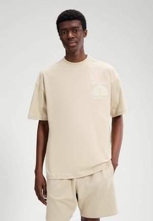 Junger Mann trägt ein beigefarbenes, oversized T-Shirt und passende Shorts, steht mit einer Hand in der Tasche vor einem schlichten Hintergrund.