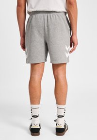 Homme portant un short de sport gris avec des logos en chevron blancs, des chaussettes blanches avec des rayures noires et des baskets noir et blanc vues de derrière.