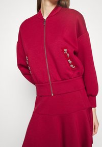 Czerwona bluza z zamkiem z przodu, ściągacze na rękawach oraz dekoracyjne zdobienia w postaci kwiatów na kieszeniach. Miękki materiał o luźnym kroju.