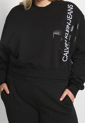 Person wearing black Calvin Klein Jeans sweatshirt and matching black pants with hands in pockets against plain background.