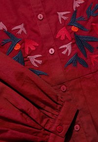 Červená košeľa s gombíkmi, ktorá má kvetinovú výšivku v ružovej, červenej a modrej farbe. Štruktúrovaná tkanina so skladmi pri leme a detailami gombíkov.