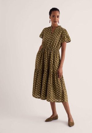Femme portant une robe midi vert olive à manches courtes avec imprimé fleuri blanc, des ballerines marron et des boucles d’oreilles rouges, debout devant un fond uni.