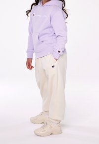 Sudadera lila con el logo "Champion" en blanco, pantalones jogger crema con logo en el bolsillo, y zapatillas beige con logo, todo en tejidos suaves y lisos.