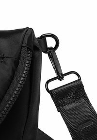 Černá nylonová taška s rohem, který obsahuje zip, nastavitelný popruh a kovové spony. Popruh má texturovaný tkaný detail.