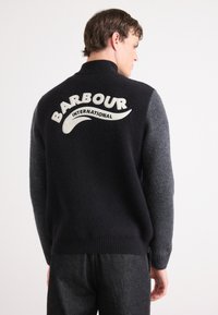 Pullover de lã azul-escuro e cinza, com um colarinho alto. Apresenta um proeminente logótipo branco "Barbour International" nas costas. Tecido de malha texturizada.