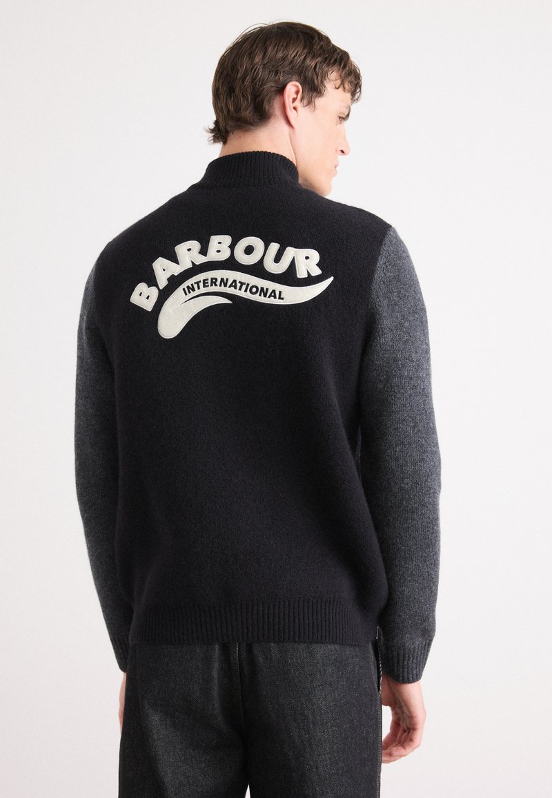 Pullover de lã azul-escuro e cinza, com um colarinho alto. Apresenta um proeminente logótipo branco "Barbour International" nas costas. Tecido de malha texturizada.