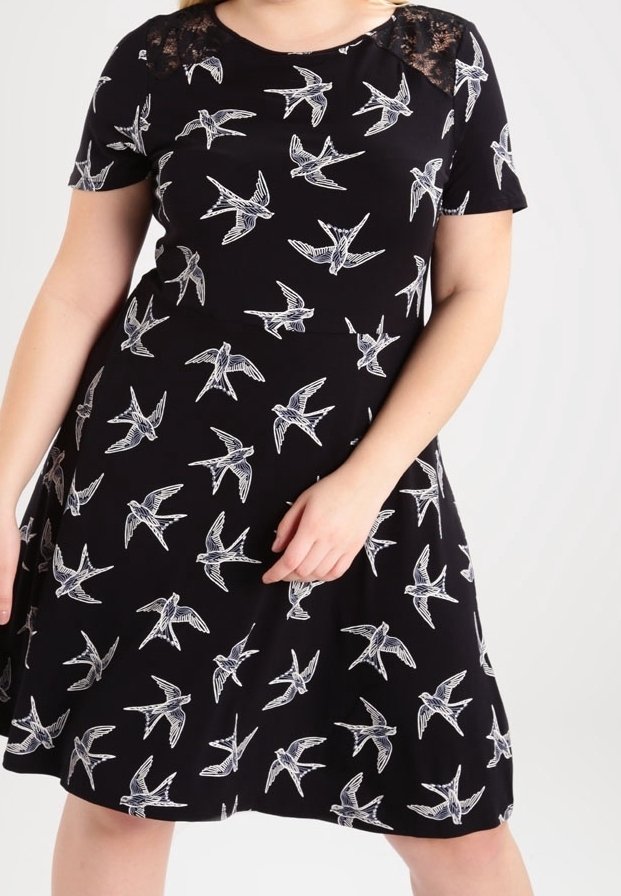 Schwarzes Kurzarmkleid mit weißem Vogelmotiv und Spitzenakzenten an den Schultern, getragen von einer Person, die vor einem neutralen Hintergrund posiert.