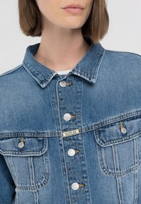 Jeansjacka i ljusblått med klassisk krage, silverknappar och två framfickor. Har märkesetikett och kontrasterande sömmar.