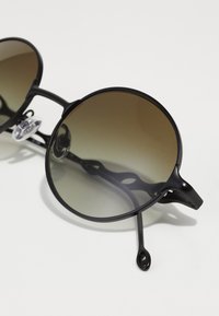 Ochelari de soare rotunzi cu ramă din metal negru, lentile brune cu un gradient și un design elegant al brațelor, care include o decupare decorativă pe tâmple.