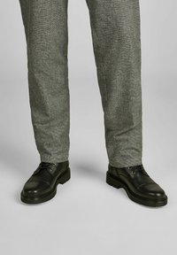 Zwarte lederen schoenen met een gestructureerde afwerking en een kaptoog, gecombineerd met lichtgrijze gestructureerde broek, beide weergegeven vanaf de taille naar beneden.