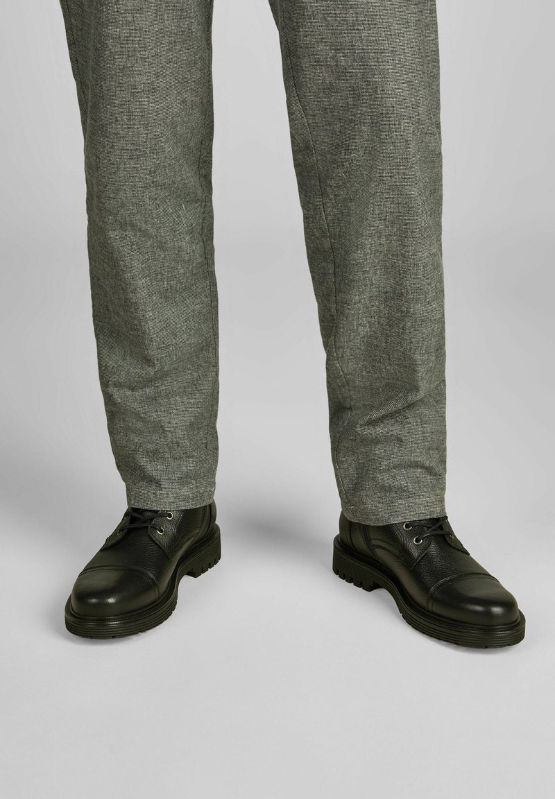 Zwarte lederen schoenen met een gestructureerde afwerking en een kaptoog, gecombineerd met lichtgrijze gestructureerde broek, beide weergegeven vanaf de taille naar beneden.