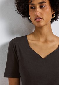 Tmavě šedé tričko s véčkovým výstřihem a krátkými rukávy. Měkká, hladká textura, minimalistický design. Zvýrazněné tónem pleti a jednoduchými zlatými kruhovými náušnicemi.