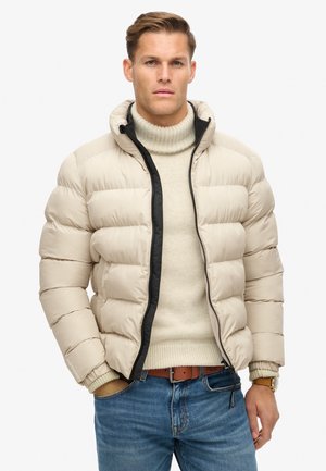 Chaqueta de plumas beige con cuello alto, cremallera negra y puños elásticos. Llevada sobre un suéter ligero y jeans, mostrando un estilo casual.