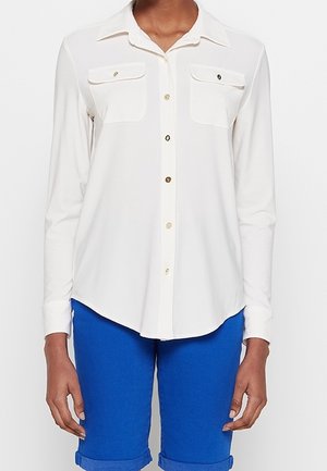 Hvid langærmet skjorte med knapper og to brystlommer, båret med lyseblå shorts, der er rullet op.
