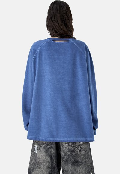 Personne aux cheveux longs et foncés portant un sweat-shirt bleu ample et un pantalon noir délavé et usé, debout de dos sur un fond uni.