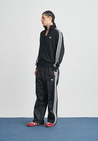 Čierna tepláková súprava z hladkej tkaniny s bielym dizajnom troch pruhov na rukávoch a nohaviciach. Červené športové tenisky zodpovedajúce súprave.