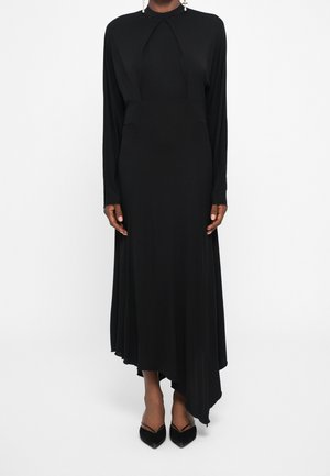 Femme portant une longue robe noire asymétrique à manches longues, associée à des talons noirs pointus et des boucles d'oreilles pendantes.