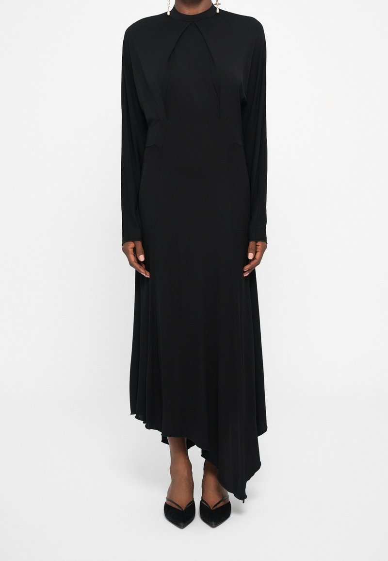 Femme portant une longue robe noire asymétrique à manches longues, associée à des talons noirs pointus et des boucles d'oreilles pendantes.