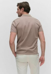Beige pikétröja med korta ärmar, har en krage och slät textur, bärs med ljusa byxor; sedd bakifrån.