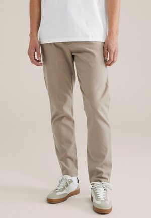 Man die beige slim-fit broek, witte sneakers met gumzolen en een wit T-shirt draagt, staand tegen een effen achtergrond.