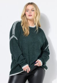 Donna bionda che indossa un oversized pullover verde scuro con rifiniture bianche e pantaloni di pelle nera, in posa contro uno sfondo bianco.