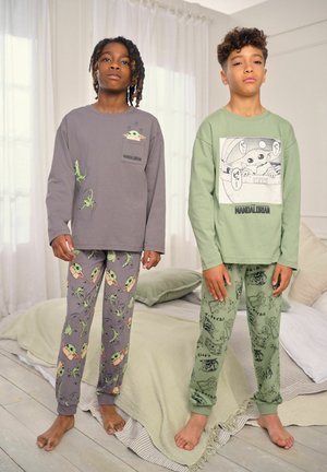 To børn står barfodet iført langærmede pyjamas med tema fra The Mandalorian og mønstrede bukser i et soveværelse.