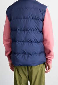 Man som bär en marinblå vadderad väst över en rosa sweatshirt och olivgröna byxor, står vänd bort mot en neutral bakgrund.