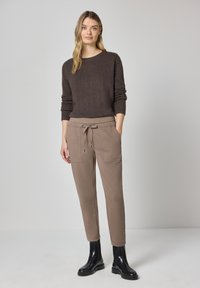 Bruine gebreide trui met lange mouwen, gecombineerd met lichtbruine taps toelopende joggingbroek met een trekkoord en twee voorzakken. Zwarte enkellaarzen.