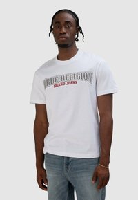 Wit katoenen T-shirt met een grafische print 'True Religion Brand Jeans' in grijs en rood. Korte mouwen en een ronde hals.