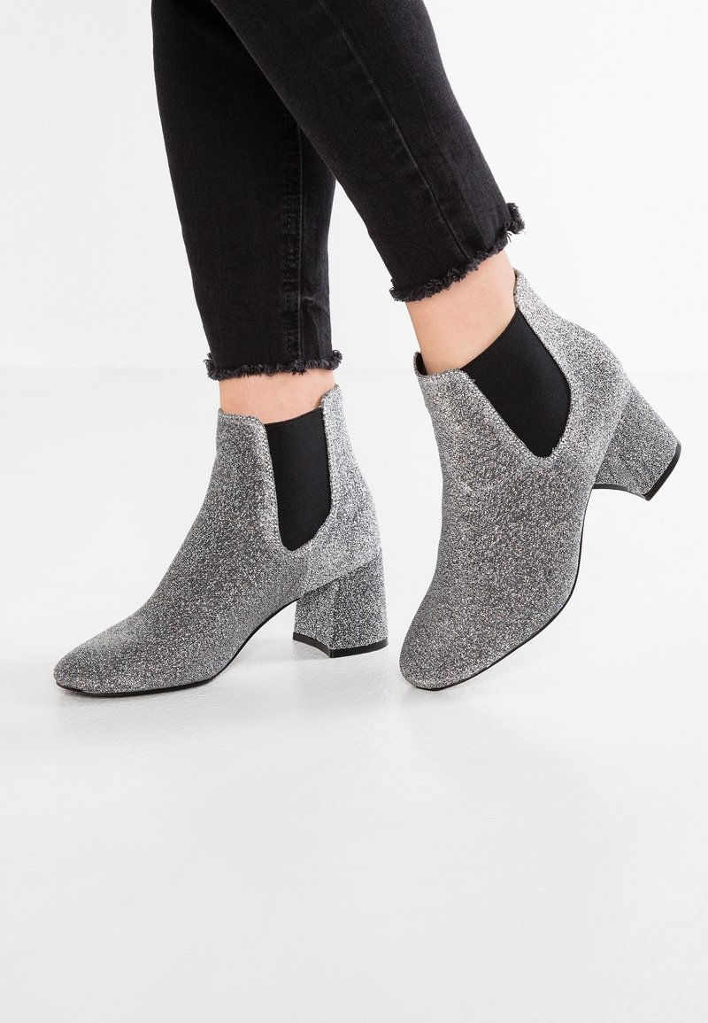Jambes en jean noir à ourlet effiloché portant des bottines argentées pailletées à talons blocs avec des panneaux latéraux élastiques noirs sur un sol blanc.