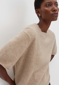 Persona di carnagione scura con capelli corti, che indossa orecchini a cerchio in argento e un maglione corto in pelliccia beige, su uno sfondo bianco semplice.