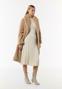 Płaszcz z sztucznego futra w kolorze camel, nałożony na beżową sukienkę swetrową z prążkowanej tkaniny. Modelka nosi jasnobeżowe botki za kostkę z szpiczastym noskiem i zamkiem błyskawicznym.