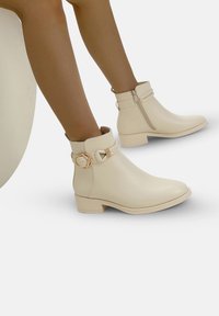 Botas de tobillo beige fabricadas en material suave, con punta redondeada, tacón bajo en bloque, cremallera lateral y una correa decorativa con un hebilla circular.