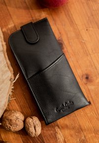 Black leather wallet with button strap on wooden surface next to walnuts and part of an apple.